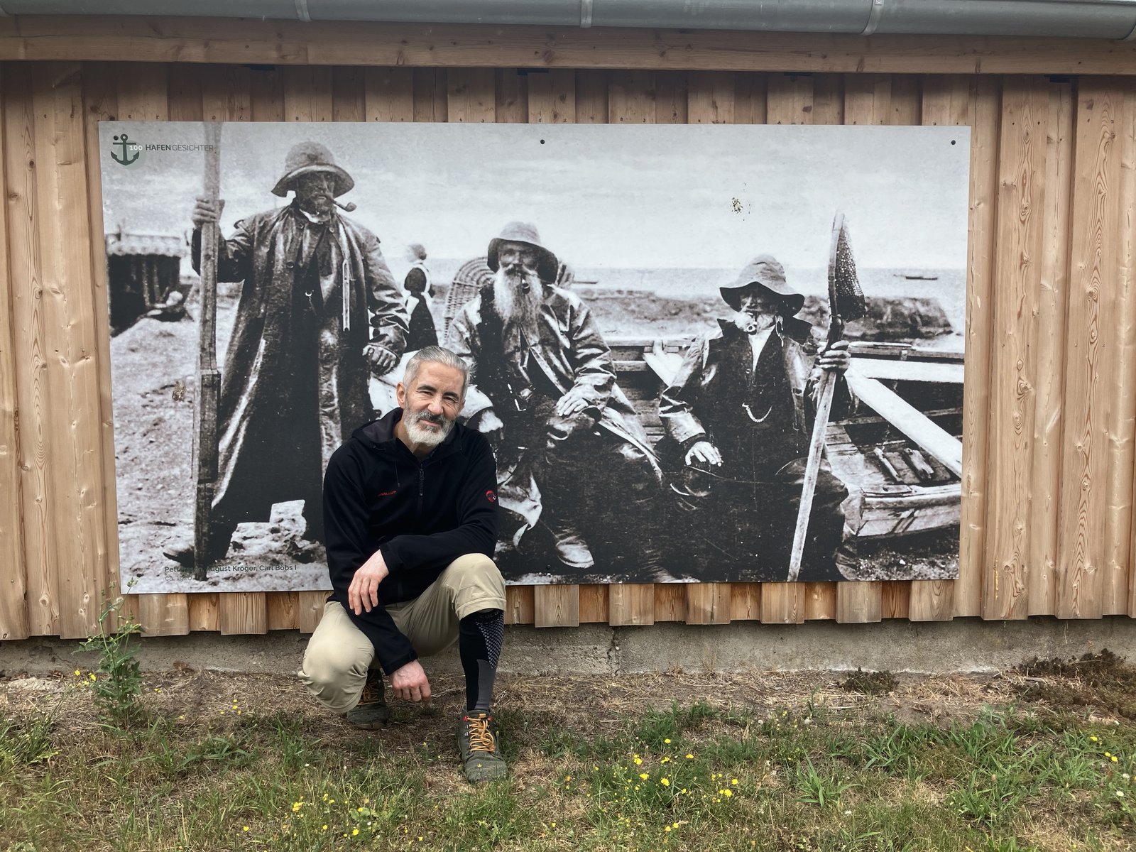 Kai Gröbner kniet vor einer großen historischen Fotografie von drei Fischern an einer Holzwand